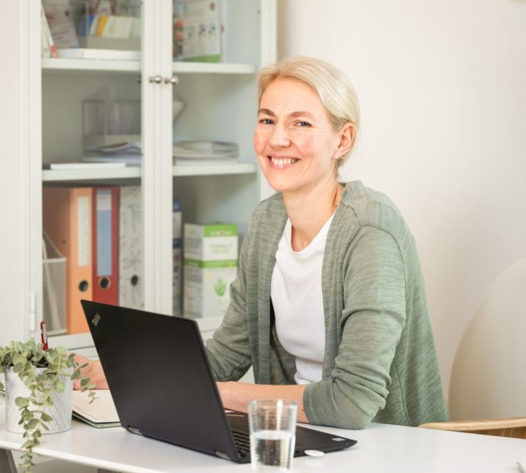 Anamnesegespräch & Behandlung Erst-Termin Lächelnde Frau mit Laptop am Tisch, Wasserglas und Pflanzen im Hintergrund.