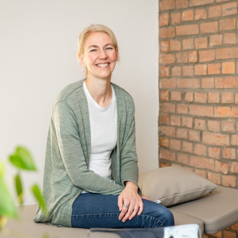 Heilpraktiker/in & Physiotherapeut/in Katrin Knüdeler Lächelnde Frau mit blonden Haaren sitzt entspannt auf einer Therapieliege vor einer Backsteinwand.